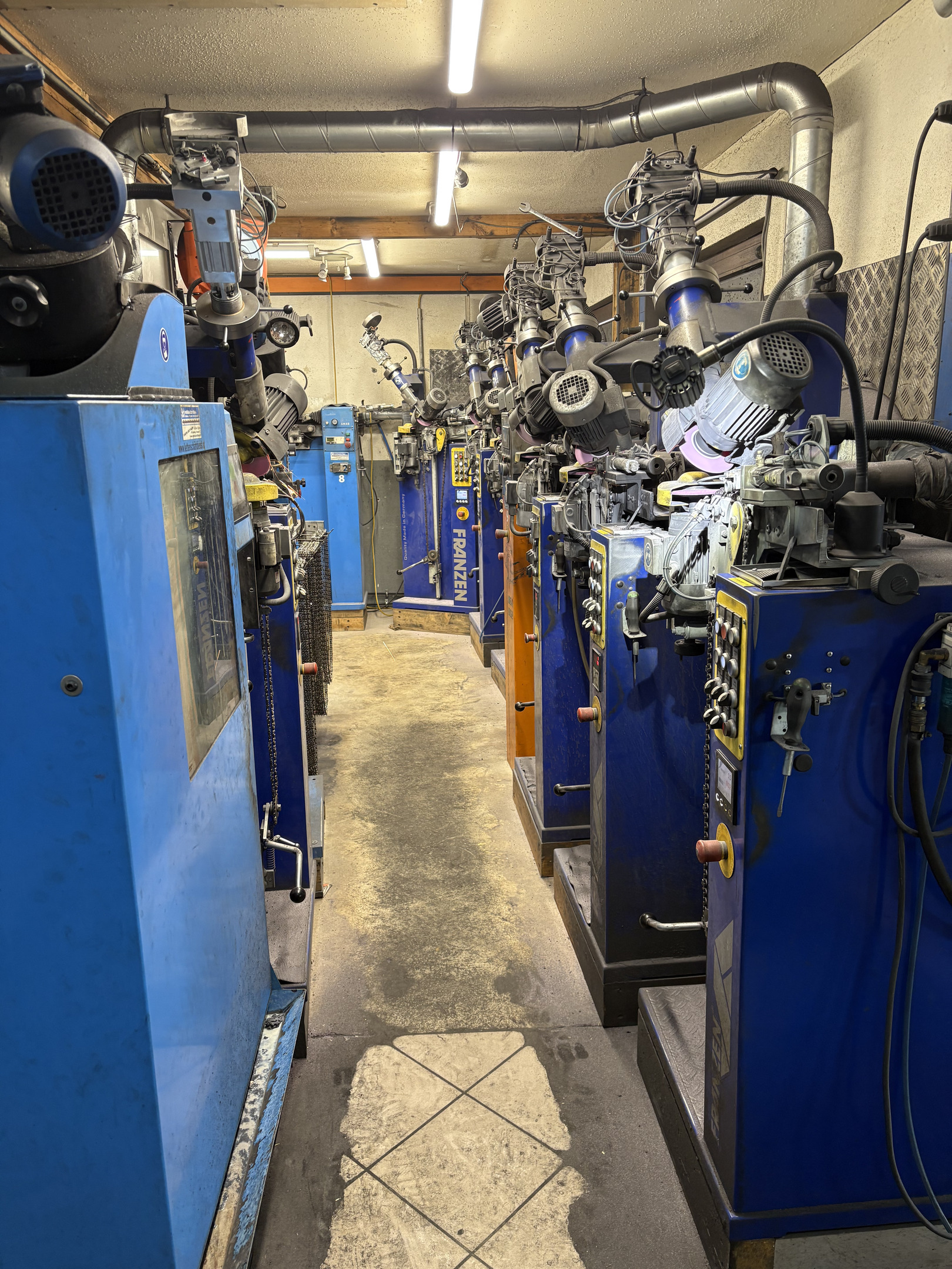 Werkstattgasse mit einer Reihe blauer Kettenschärfmaschinen, links und rechts Schleifaggregate mit Motorsägeketten